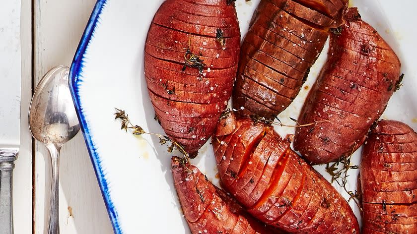 roasted hasselback sweet potatoes on a white serving plate with thyme and serving utensiles