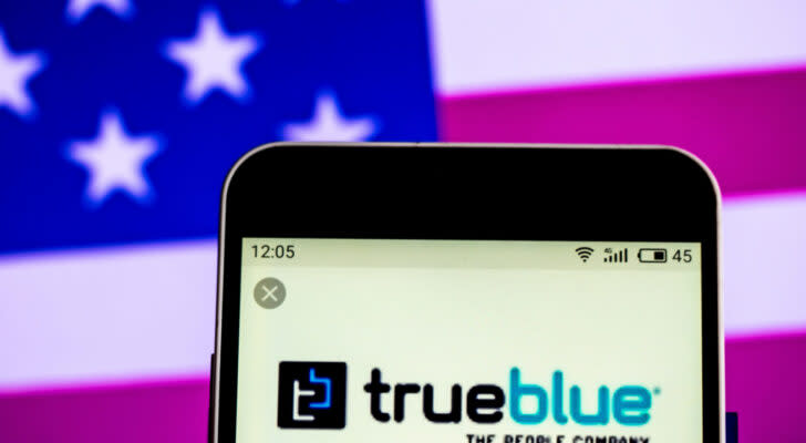 TBI stock: the TrueBlue logo on a phone in front of an american flag background