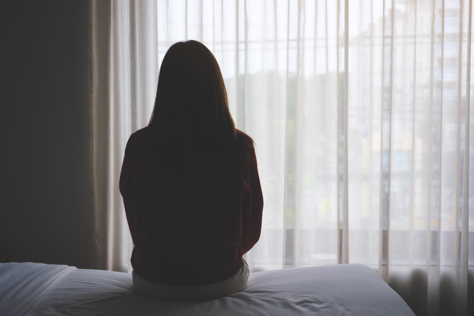 A person sits on a bed, facing a window with sheer curtains, appearing contemplative