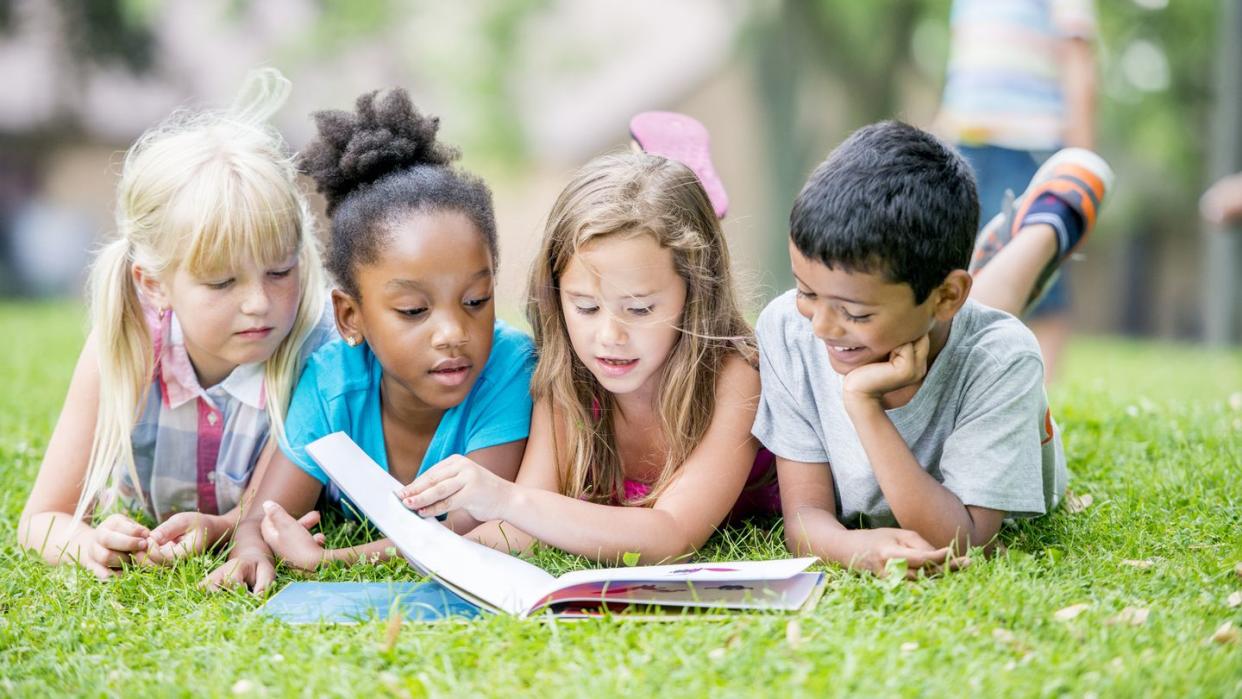 four kids laying on their bellies in the grass and reading a book together