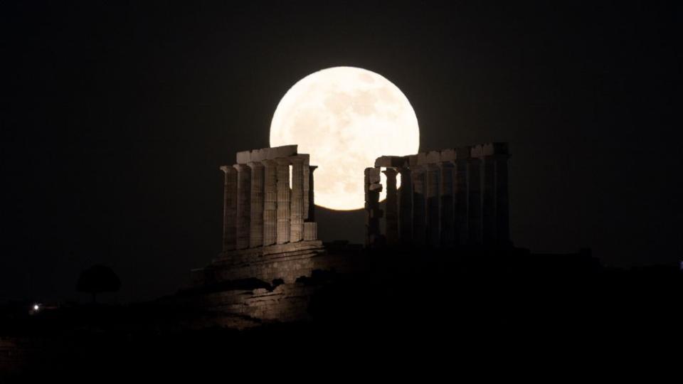 a large bright moon is seen behind the ruins of the Temple of Poseidon where several large columns rise high into the sky.