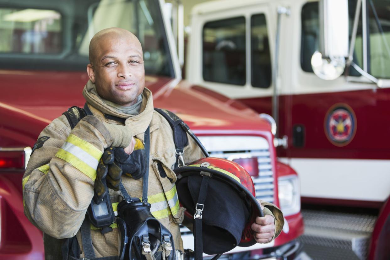 Portrait of an African American fireman standing in front of a fire engine parked at the station.  He is serious and confident, wearing protective suit, holding gloves and a helmet.  He is ready to respond to an emergency.