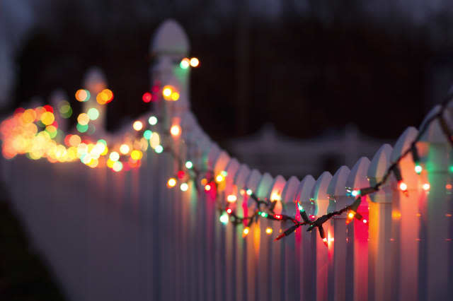 a string of multi-colored christmas lights are strung across a white plastic fence.  their reflection is cast upon the reflectiv