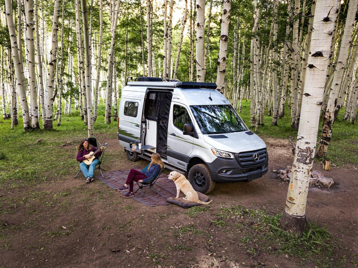 A Winnebago Revel among birch trees with two people and a dog sitting outside.