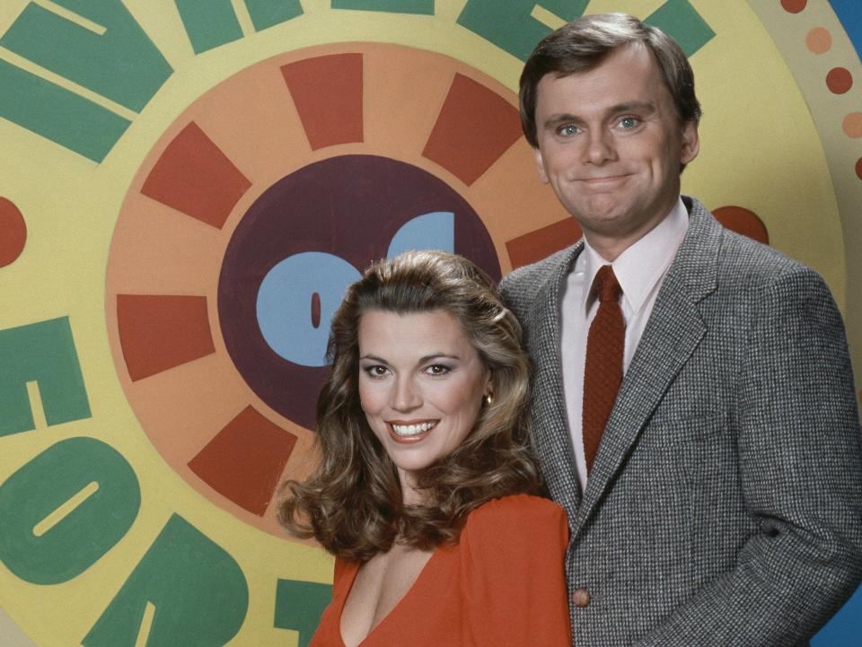 Vanna White wearing a red top, Pat Sajak holding her arm waring a suit with Wheel of Fortune logo behind them