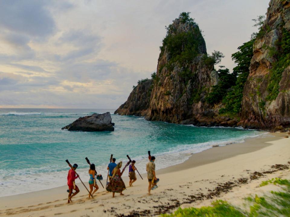 six survivor players carrying their torches to tribal council against a tropical, ocean view