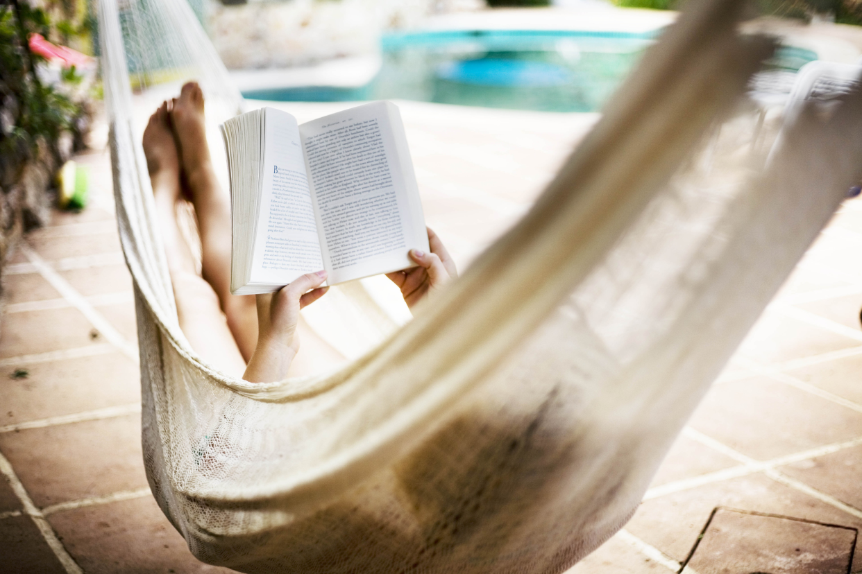 Woman reading a book while laying in a beige hammock, selective focus of book, on a patio with a swimming pool and plants blurred in the background