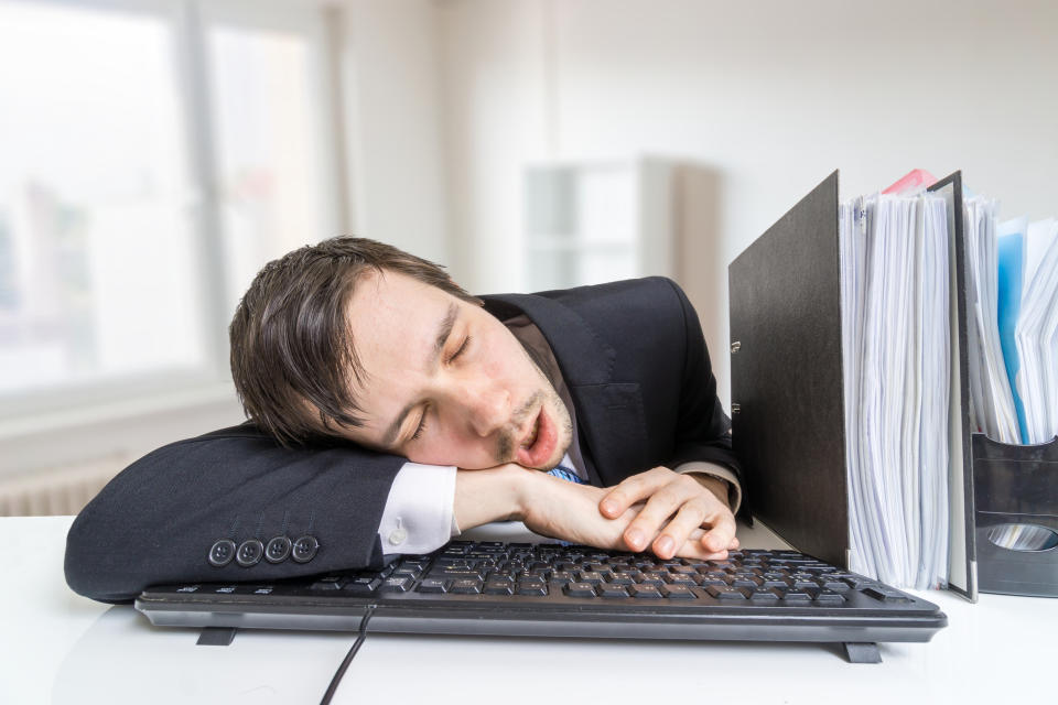 A man in a suit is asleep at his desk, resting on a keyboard with a file organizer beside him, suggesting exhaustion or overwork in a professional setting