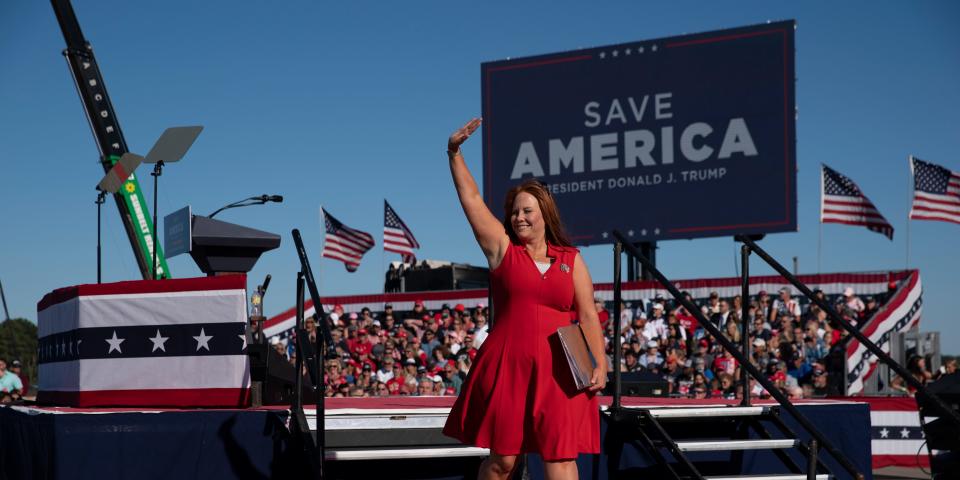Sandy Smith wears a red dress and waves in front of a "Save America" sign.