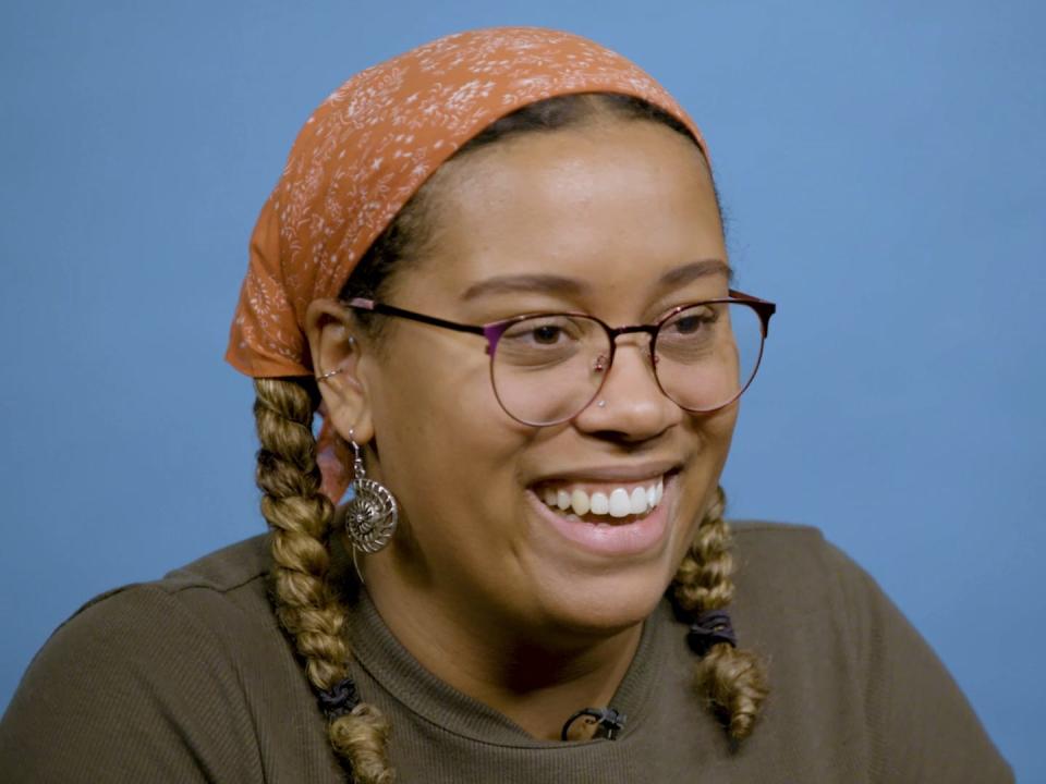 A still shows Anima Webber-Shultz smiling on a blue background. She is a dark skinned woman wearing glasses. She is wearing an olive green top and an orange bandana wrapped around her head. Her hair is braided in two pigtails on either side of her face.