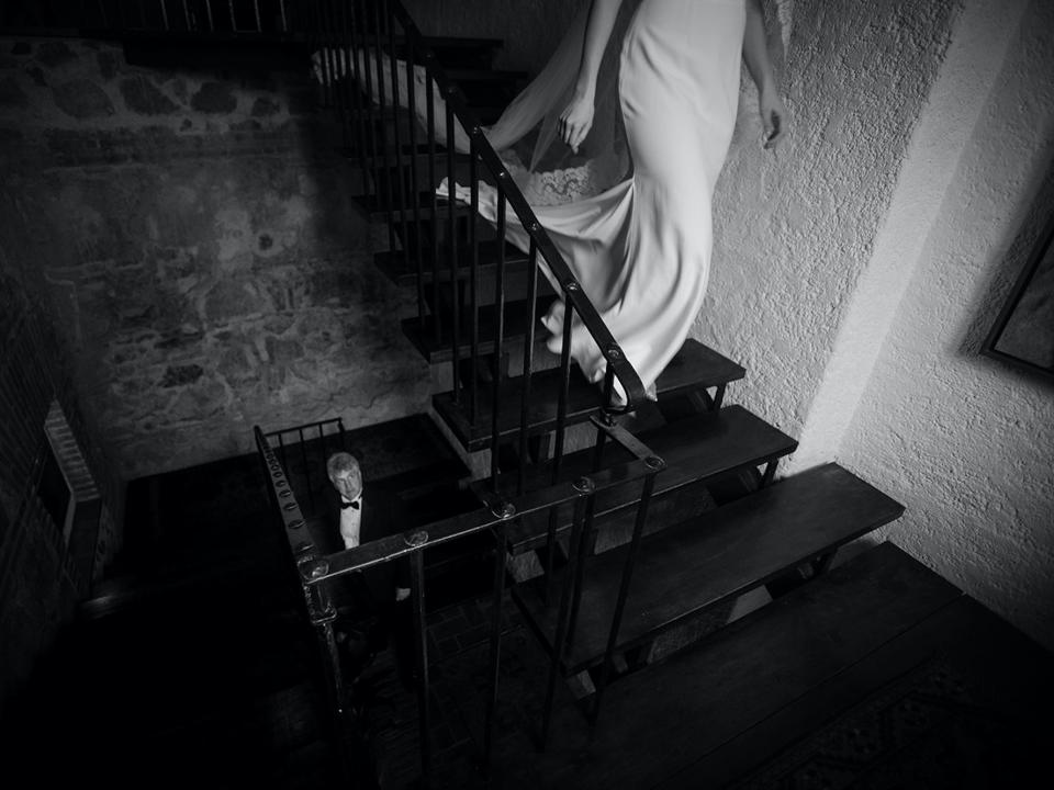 The lower half of a bride's body is visible walking down a set of stairs as her father looks up at her from a lower floor.