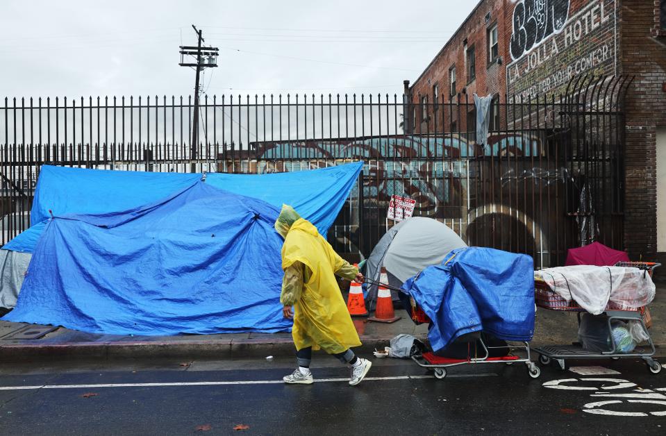 A person walks with carts in the rain near an encampment of unhoused people in Skid Row in Los Angeles on February 6, 2024.