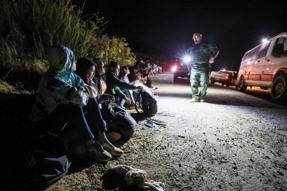 A man shines a flashlight on a line of people sitting on the ground.