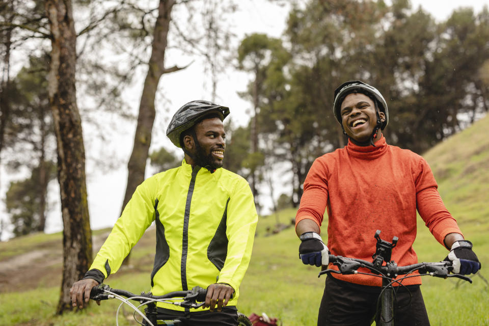 Two men, one wearing a yellow neon jacket and the other wearing a bright orange long-sleeve turtleneck top, smile as they ride bikes together