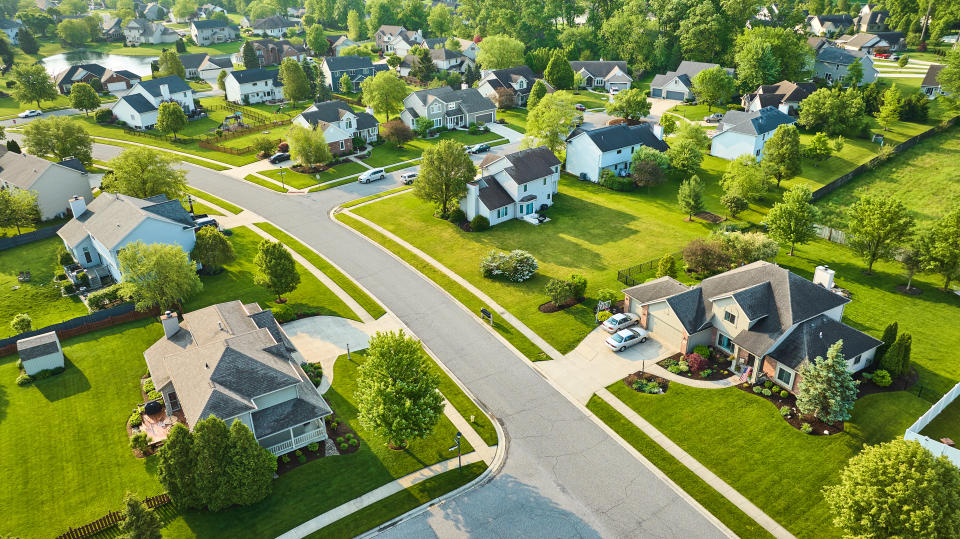 Aerial view of a suburban neighborhood with neatly arranged houses, well-maintained lawns, and tree-lined streets. No persons are visible in the image