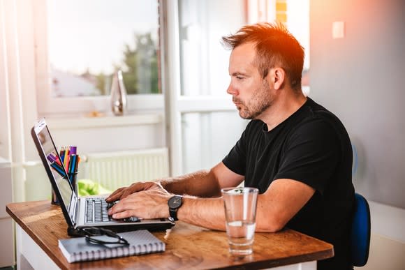 Man typing on a laptop at a small desk next to a window