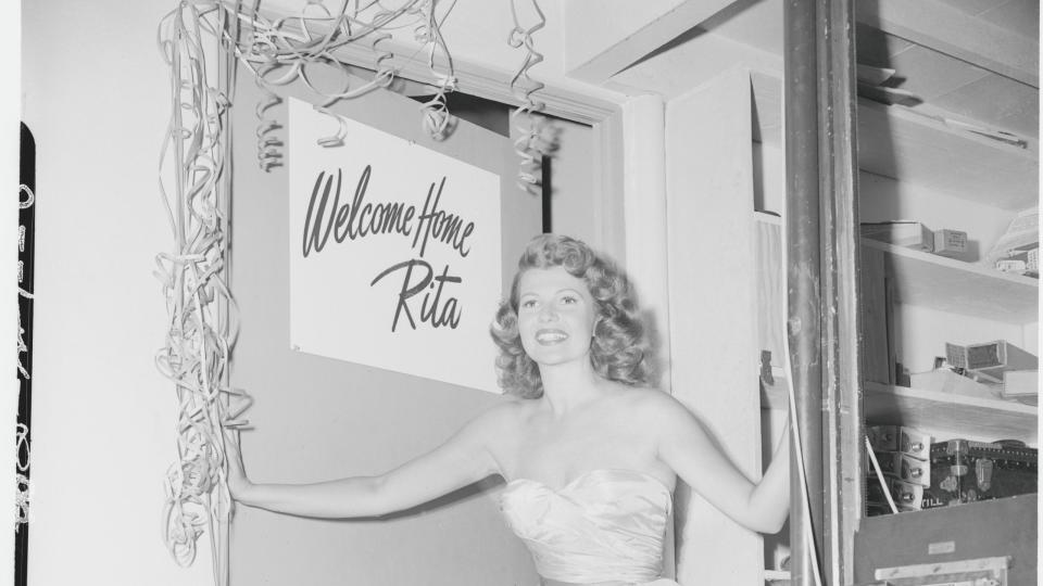 rita hayworth stands in the door way of a dressing room, she wears a strapless ballgown and a sign on the open door behind her says welcome home rita