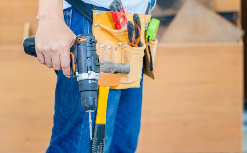 A worker in jeans wears a tool belt and holds a drill in one hand. 