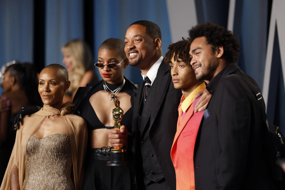 Group of people on red carpet, two men in suits, one holding an award, flanked by a woman in a sleeveless dress and one with sunglasses