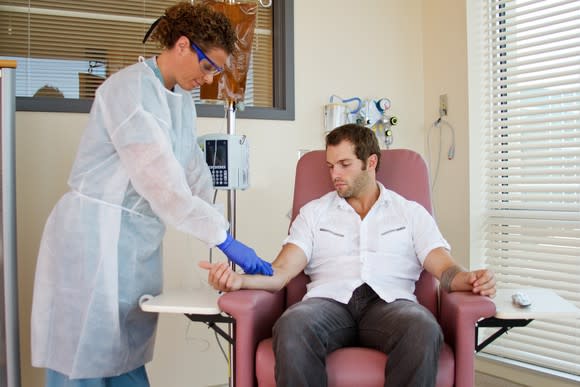 A female technician in a lab coat standing over a seated male patient and preparing him for an infusion