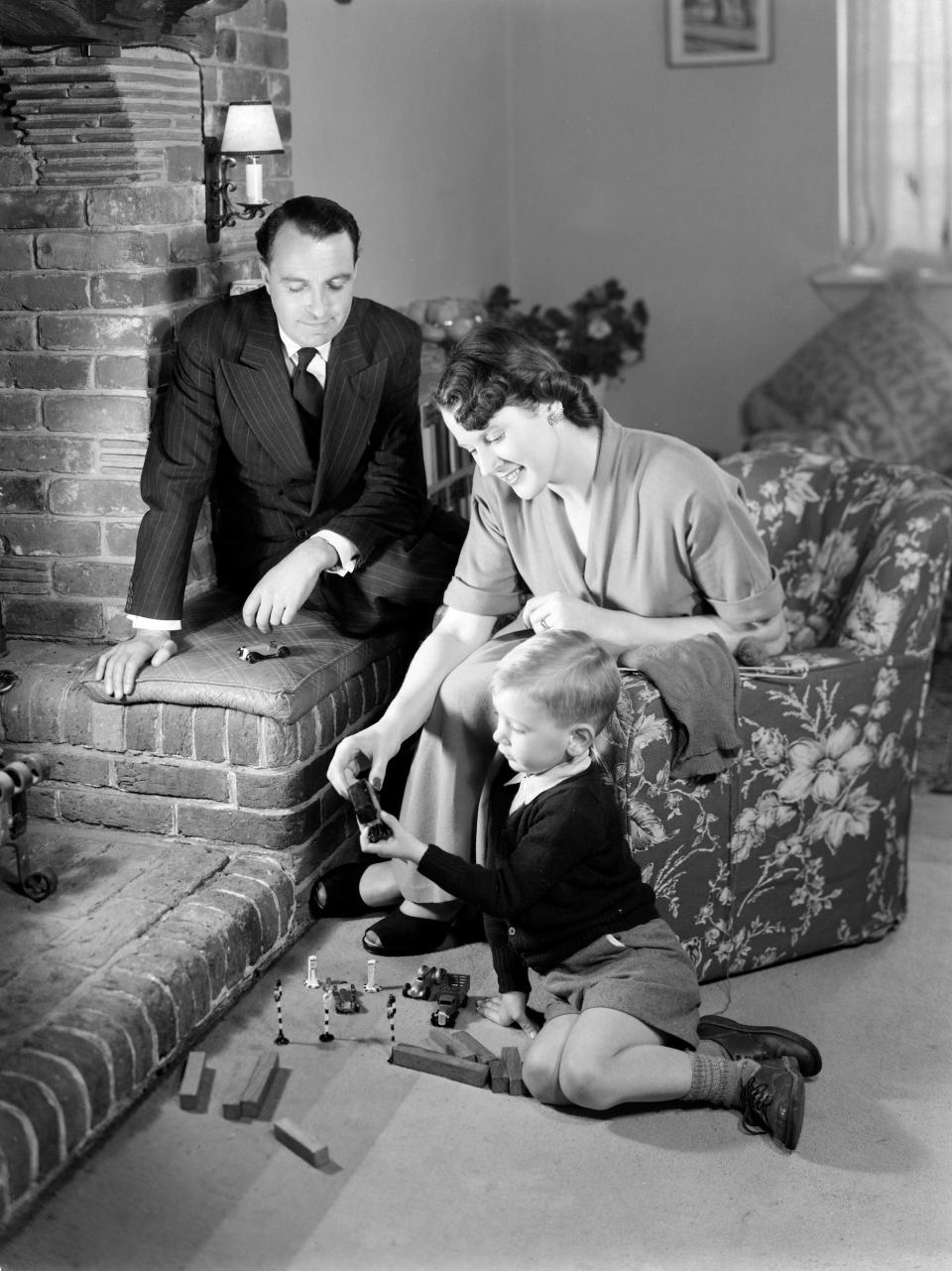 A photograph of a family relaxing in front of the fire, with parents looking on as their son plays on the floor in front of the fireplace.