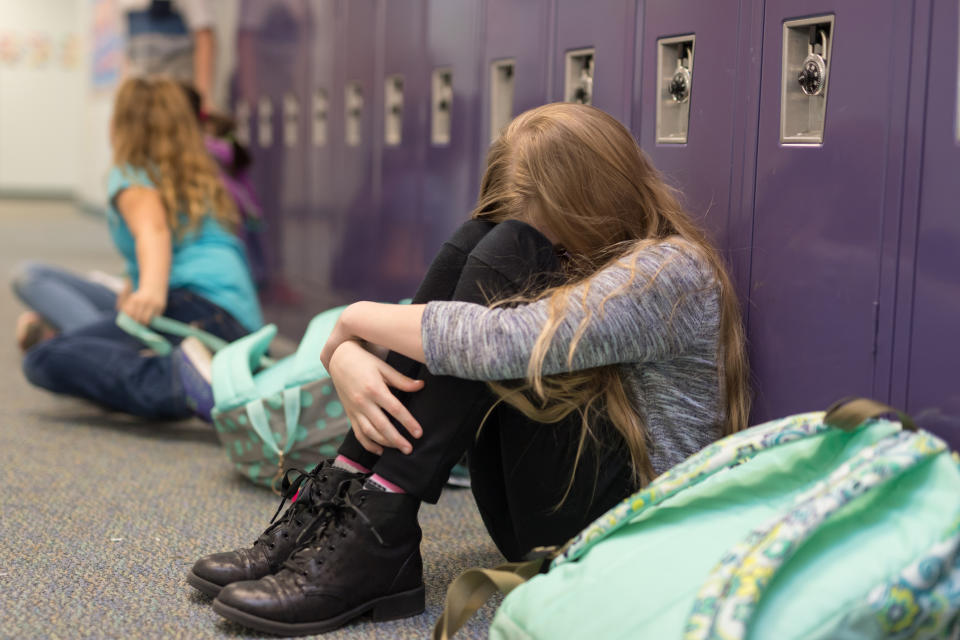 A girl sits with her head down against school lockers, hugging her knees, while another girl sits nearby, both with backpacks