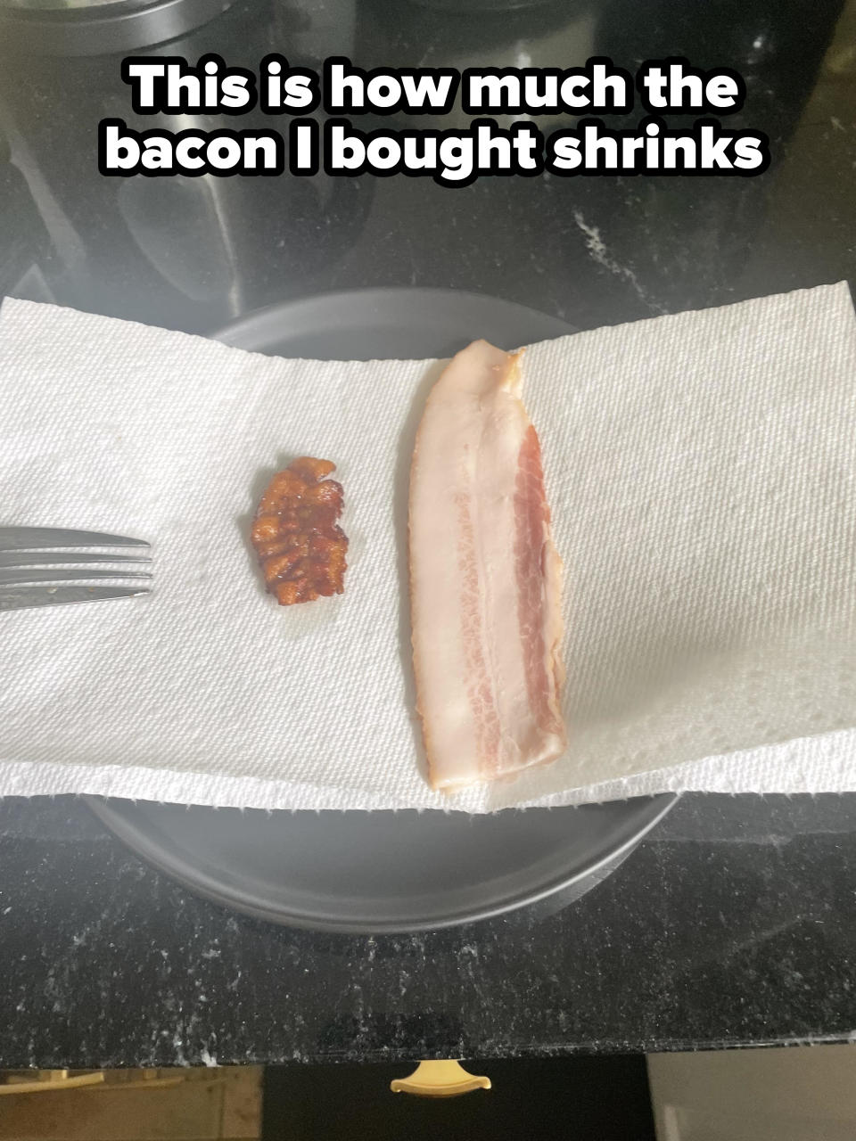 A strip of uncooked bacon and a small piece of cooked bacon on a paper towel with a fork on the left, placed on a dark kitchen countertop