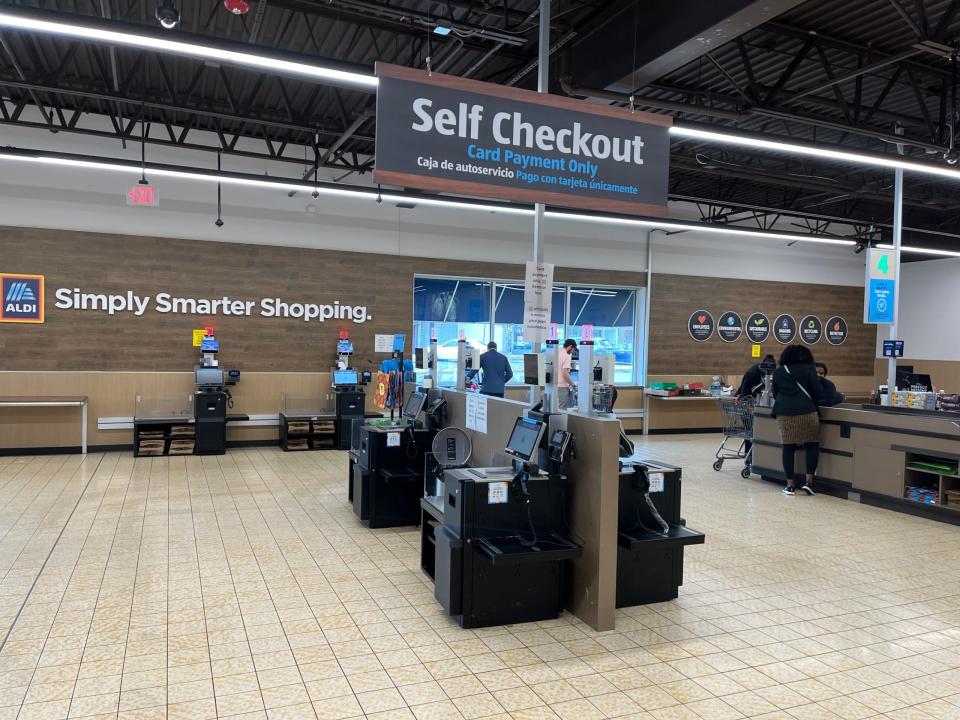 Vor einem Aldi-Markt in Maryland steht eine Reihe von Selbstbedienungskassen. Auf einem Schild über ihnen steht: "Self Checkout, nur Kartenzahlung", gefolgt von der gleichen Botschaft in Spanisch. An der Wand hinter den Kassen sind das Aldi-Logo und der Slogan "Simply Smarter Shopping" zu sehen.