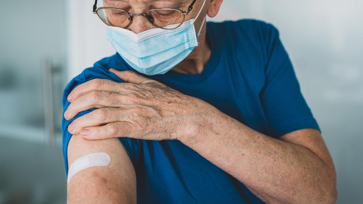  older man wearing  a blue surgical mask looks down at his arm, which bears a bandage as if he's just gotten an injection 