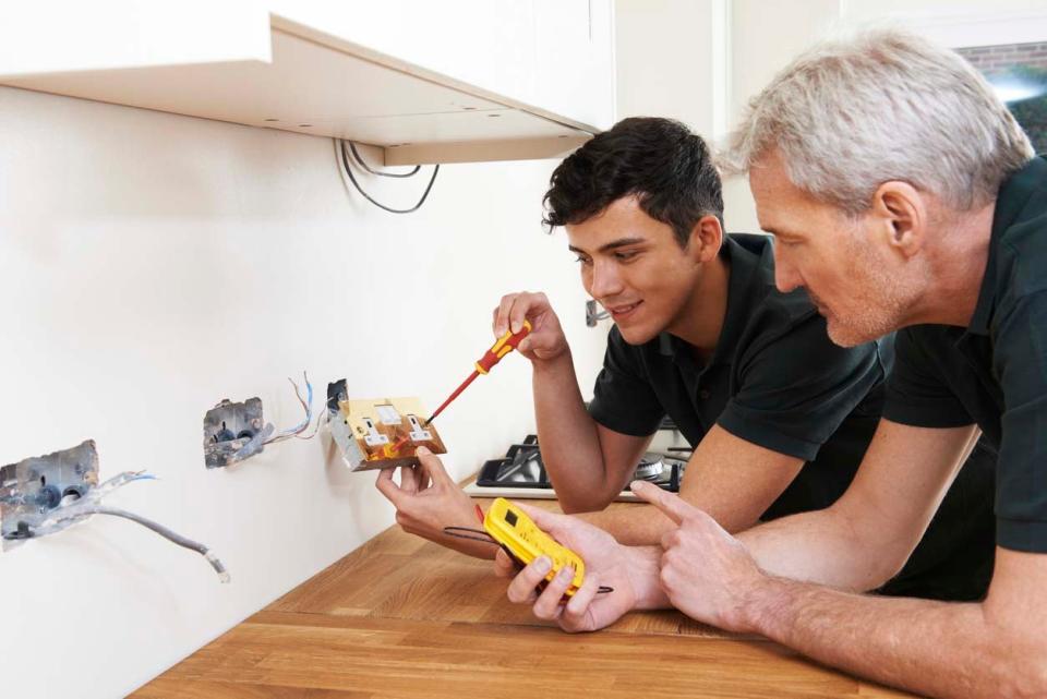 Two men uses tools to assess electrical wires protruding from a white wall.