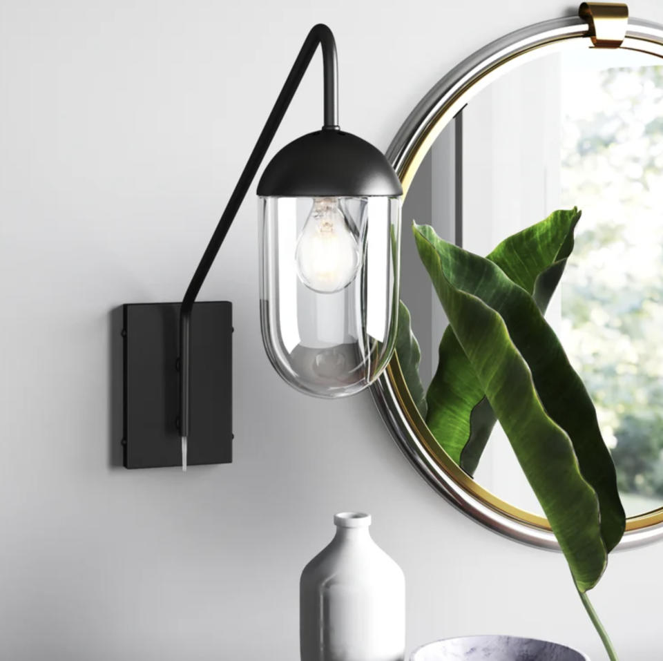 Wall-mounted light fixture with a clear shade next to a round mirror and green plant leaf