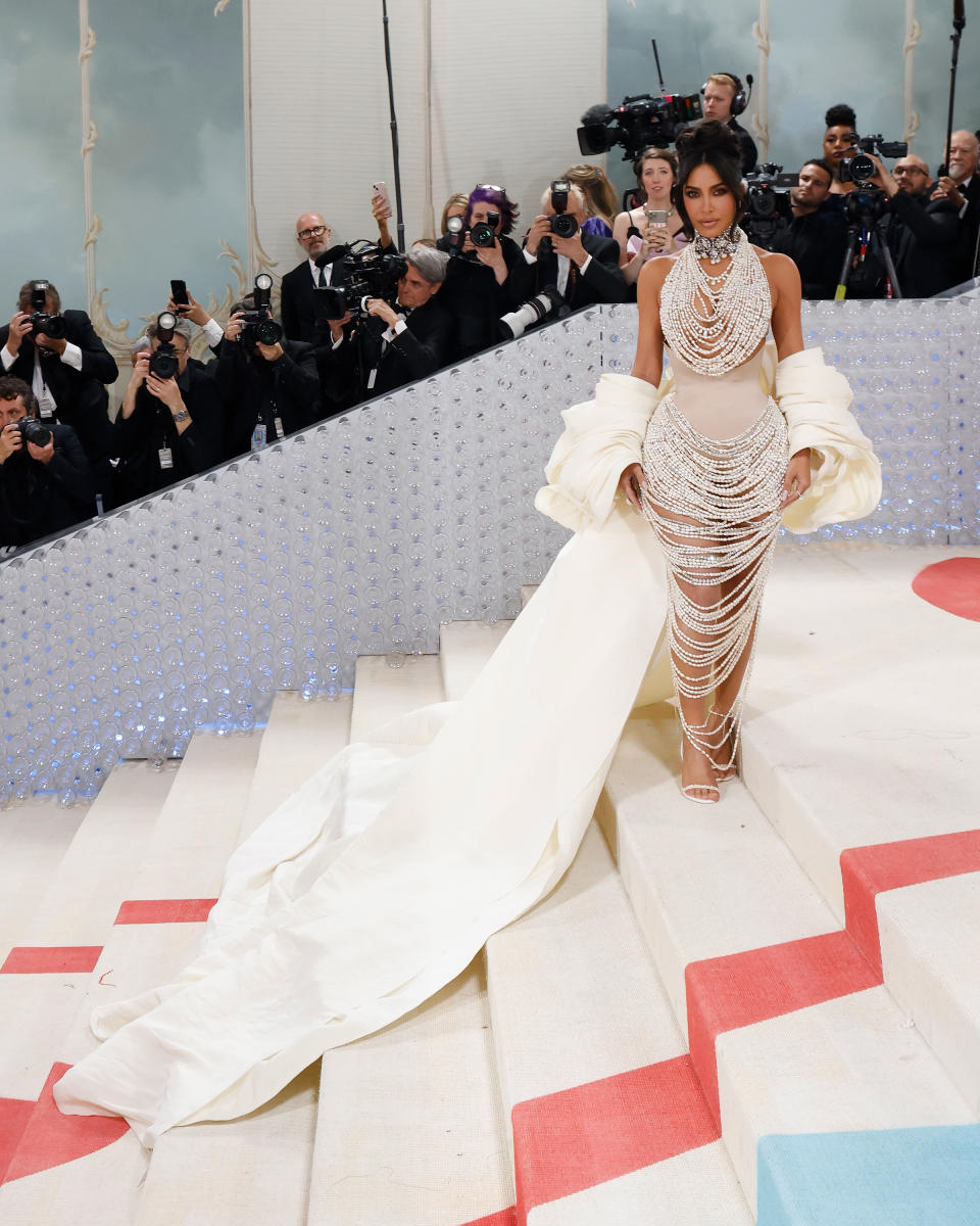 Celebrity in a beaded dress with a large ruffled train and statement necklace, posing on the event's carpet with photographers in the background