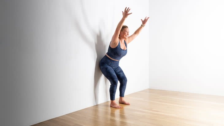 A woman with blonde hair wearing blue tights practices Utkatasana (Chair Pose) with her hips against a white wall,.