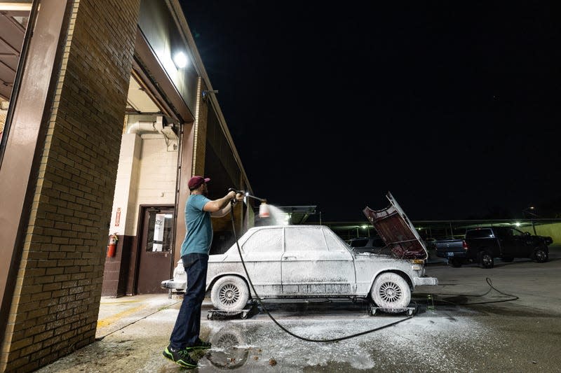 nighttime photo of the 1974 bmw 2002tii parked outside a large garage. the car is covered top to bottom in soap suds as the owner sprays the car with a pressure washer.