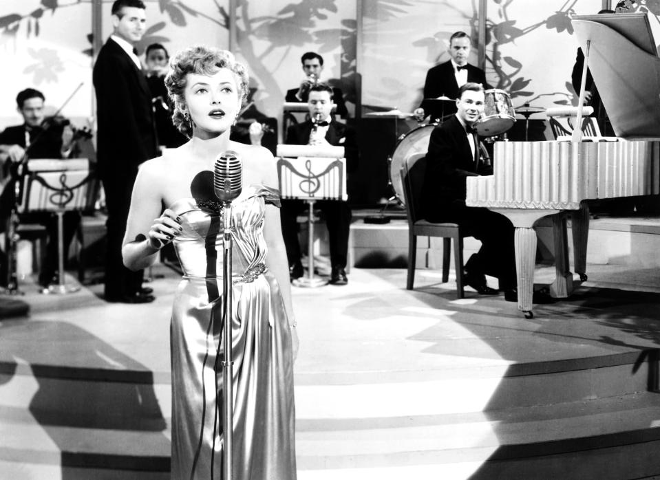 Kitty in an elegant dress sings into a vintage microphone on stage, with a pianist and band playing in the background