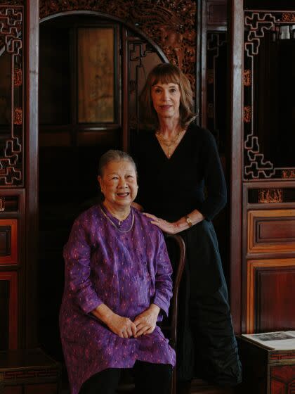 Two women, one seated and one standing, pose inside an antiques store.