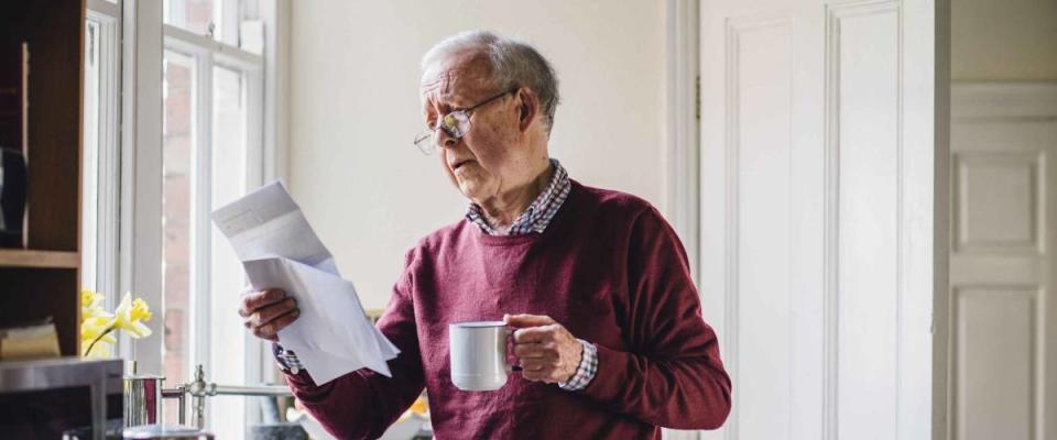 Senior man is standing in the kitchen of his home with bills in one hand and a cup of tea in the other. He has a worried expression on his face.