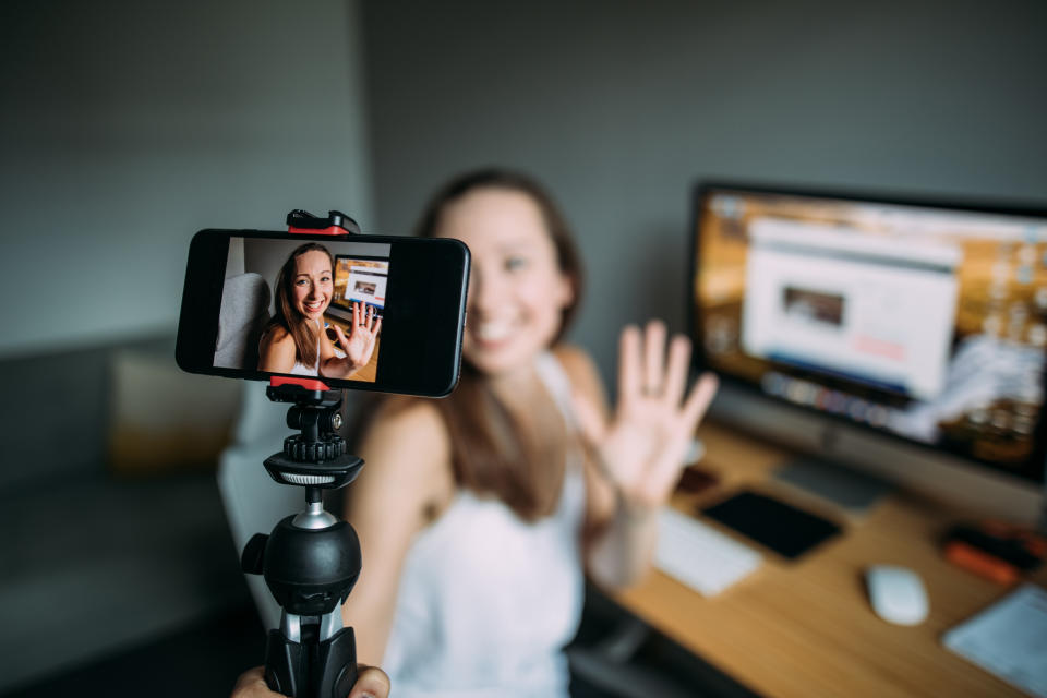 Woman recording a video at a desk with a smartphone mounted on a tripod. She's smiling and waving at the camera