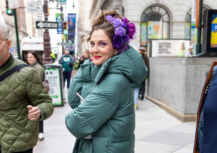 Drew Barrymore with purple flowers in hair, wearing a puffer jacket on a city street