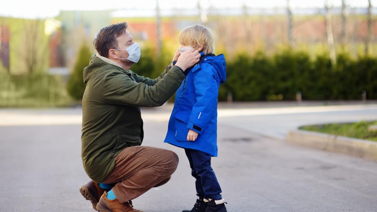 Mature man wearing a protective mask puts a face mask on a his son in airport, supermarket or other public place.