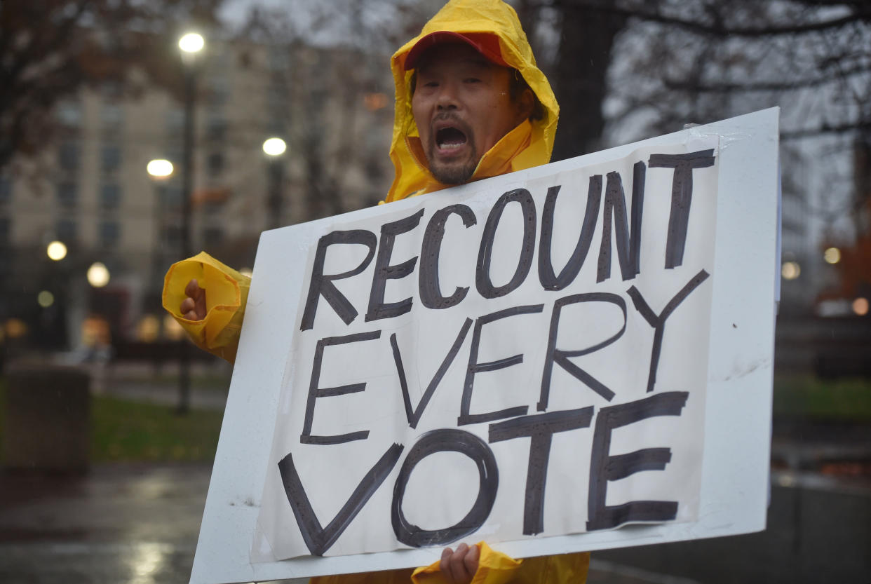 A protester in a yellow slicker holds a placard that says: Recount Every Vote.