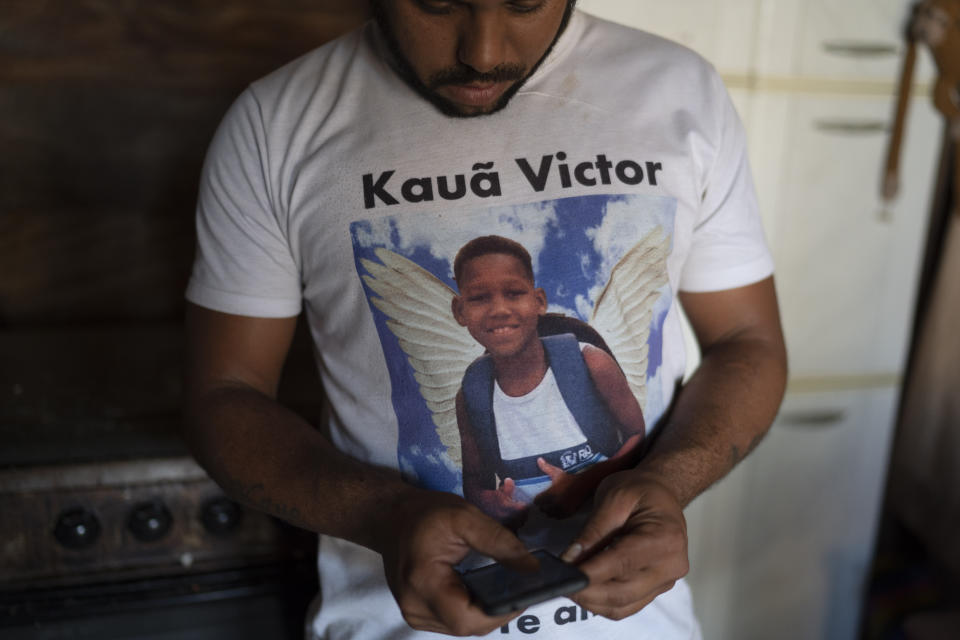 In this June 20, 2019 photo Jocely Rozario Junior wears a T-shirt emblazoned with the image of his son Kaua, who was killed by a stray bullet, as he uses a cellphone inside his home at the Vila Alianca slum in Rio de Janeiro, Brazil. "My son wasn't an adult or a trafficker. He wasn't anything like that, but he lived with fear every time he saw the police," Rozário Junior said. "I can't be quiet because if so, Kauã would be just one more statistic." (AP Photo/Leo Correa)