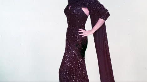 rita hayworth smiles and poses for a photo in a sparkling dark purple evening gown and matching shawl, she wears black high heels, one hand is on her hip and one hand rests on top of her head