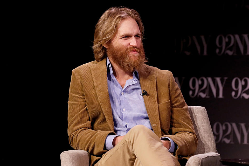 Wyatt Russell is sat on stage on a beige chair wearing a brown suit jacket, blue shirt and beige pants. 