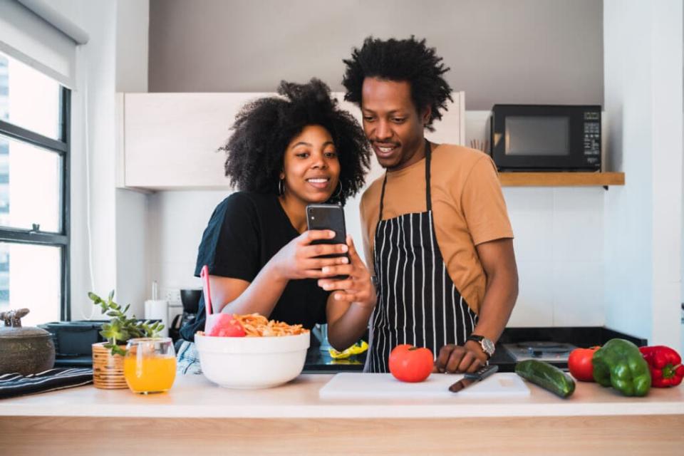 Portrait of young afro couple cooking together and using mobile phone in the kitchen at home. Relationship, cook and lifestyle concept. (Photo: Adobe Stock)