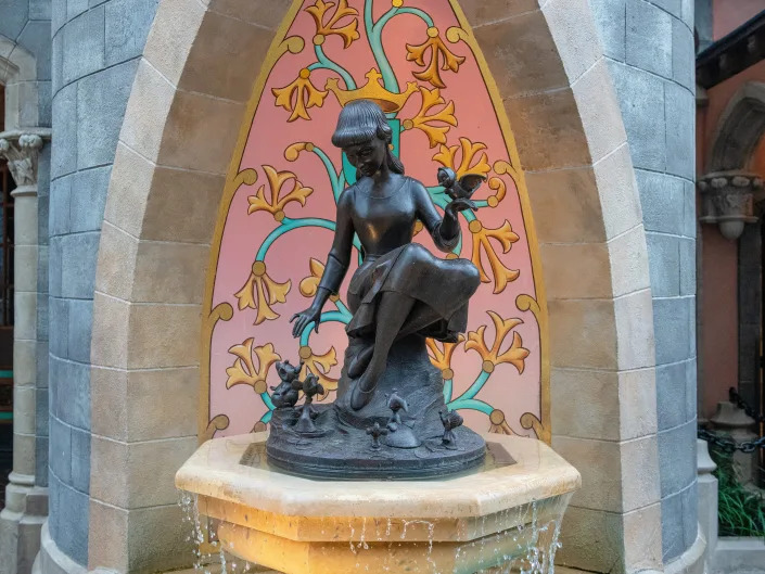 Dark metal statue of Cinderella holding a bird and reaching down to mice. It sits in front of a pink and yellow mural of flowers and a crown.
