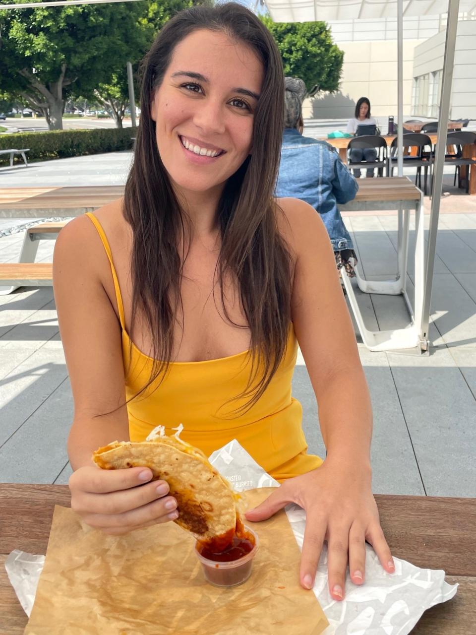 Insider's reporter wearing a yellow dress while sitting at a bench outside and holding up Taco Bell's Grilled Cheese Dipping Taco