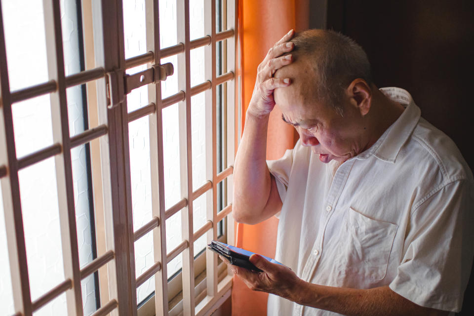 Stressed senior Asian man pressing his head, while looking at his phone, illustrating a story on Shopee improving in anti-scam rating.