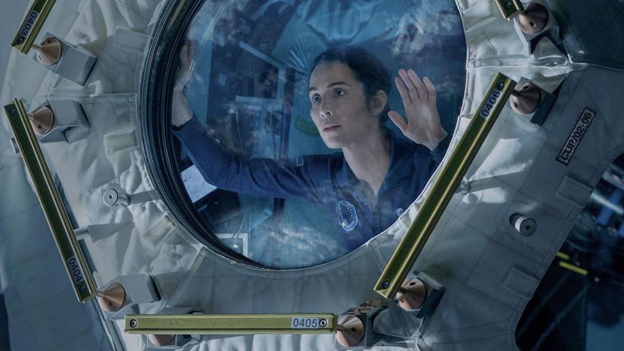  A woman in a blue flight suit looks through a glass hatch in a cramped laboratory filled with wires and screens. 