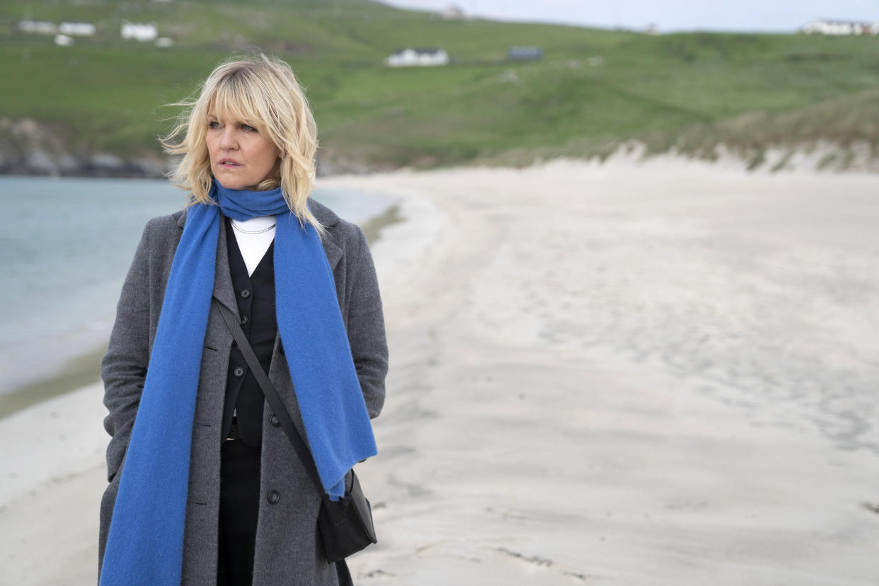  Ashley Jensen as DI Ruth Calder in Shetland. She is standing on the beach wearing a warm grey cashmere coat, with an electric blue scarf wrapped around her neck with the ends flapping in the end. Ruth's body is facing the camera but her face is looking off to the side, out to sea. 